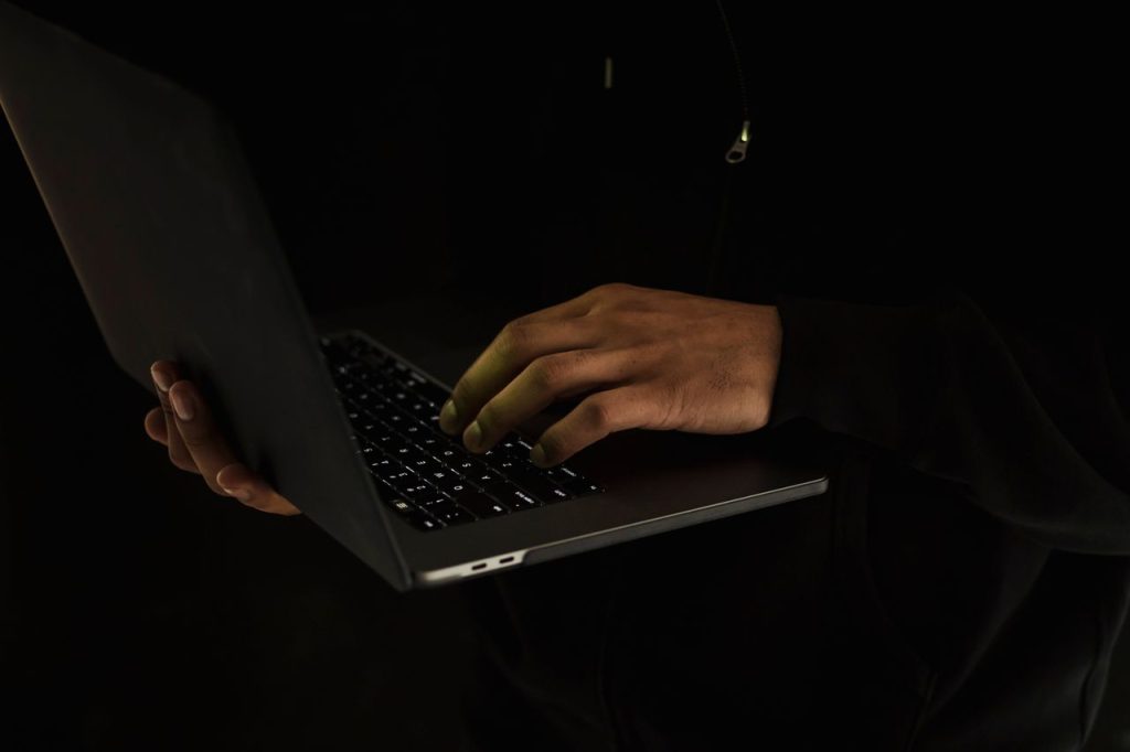 Crop anonymous male wearing black outfit standing with netbook on hand and surfing internet in dark room