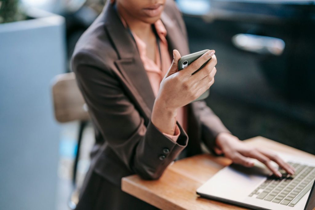 High angle of crop African American female employee wearing formal clothes typing on laptop while checking message on cellphone