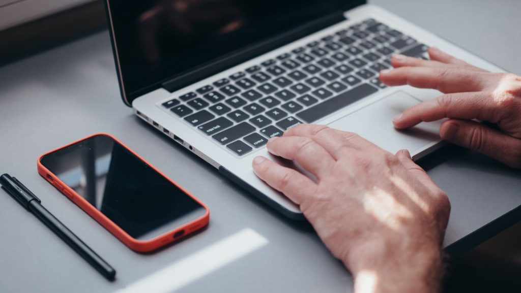 pexels photo 7054508 7054508 Hands typing on a laptop with a smartphone and pen on the side, perfect for tech and work themes.